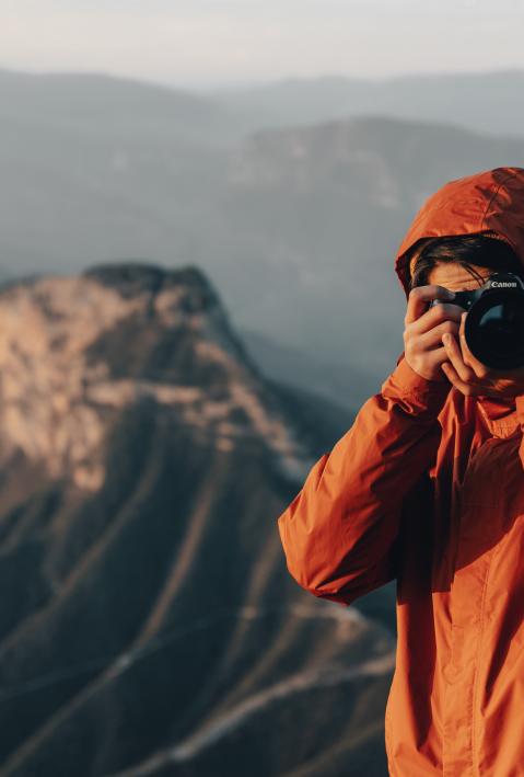 Ein Mann steht auf einem Berggipfel und schießt ein Foto und schaut direkt in die Kamera dabei. 