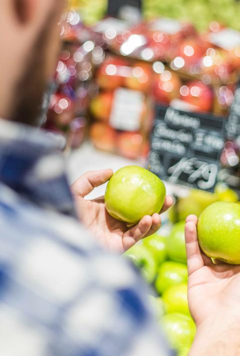 A man shopping, he looks at two green apples in his hands. 