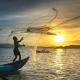 Eine Peron auf einem Fischerboot wirft Fischernetze aus - im Hintergrund ist der Sonnenaufgang. 